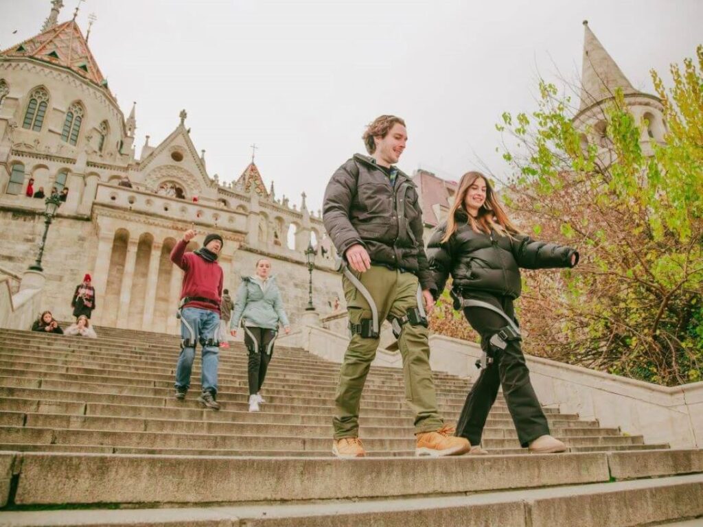 Four people wearing Hypershell exoskeletons walking down the steps in Buda Castle during a walking tour in Budapest, Hungary.